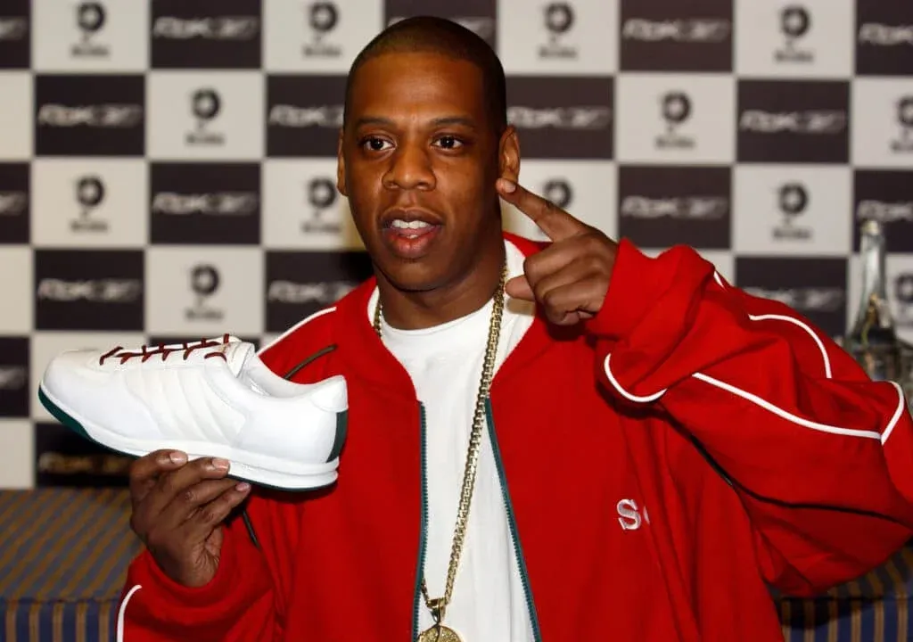 A person in a red jacket holding a white sneaker with red laces, with a patterned backdrop behind.