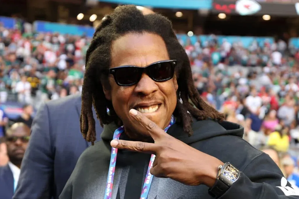 A person wearing sunglasses and a dark hoodie flashes a peace sign in front of a blurred stadium crowd.