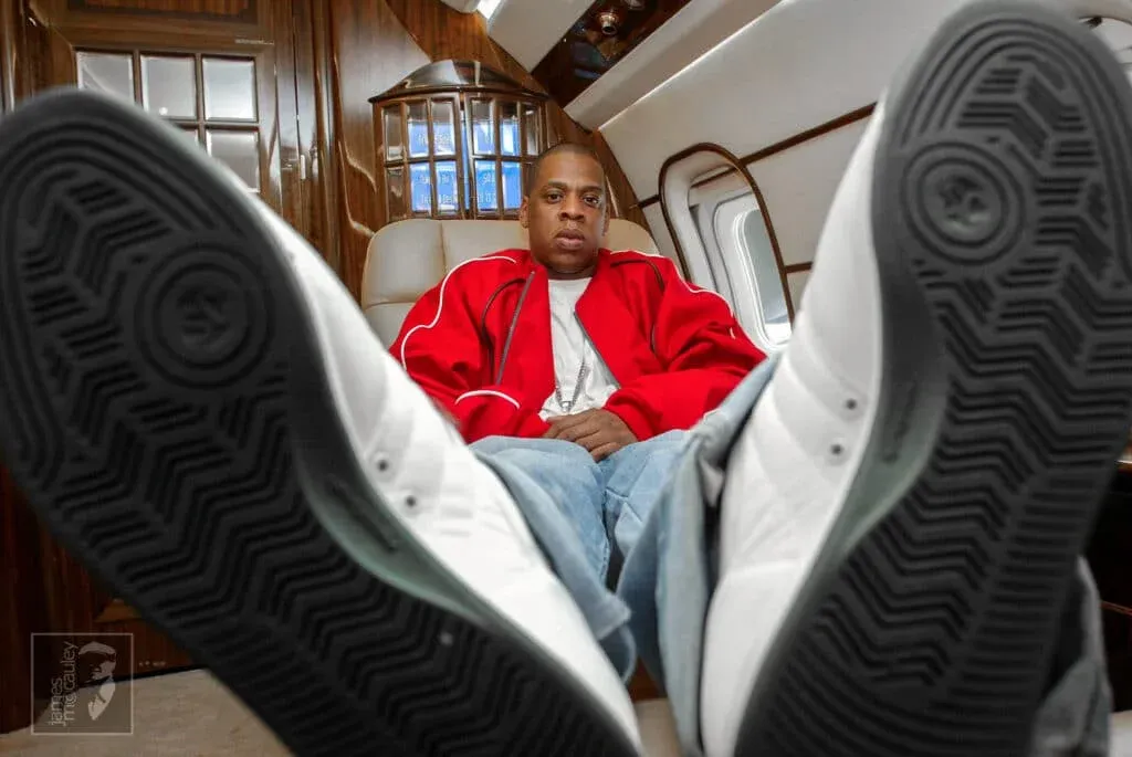 A person wearing a red jacket and jeans is seated inside a private jet, with the camera focused on the soles of their white sneakers, which are prominently displayed in the foreground.