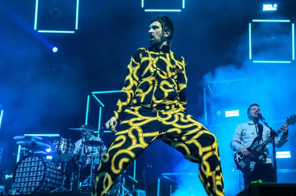 A performer in a vibrant, patterned yellow and black outfit sings energetically on stage, with a drummer and bassist in the background under blue stage lighting.