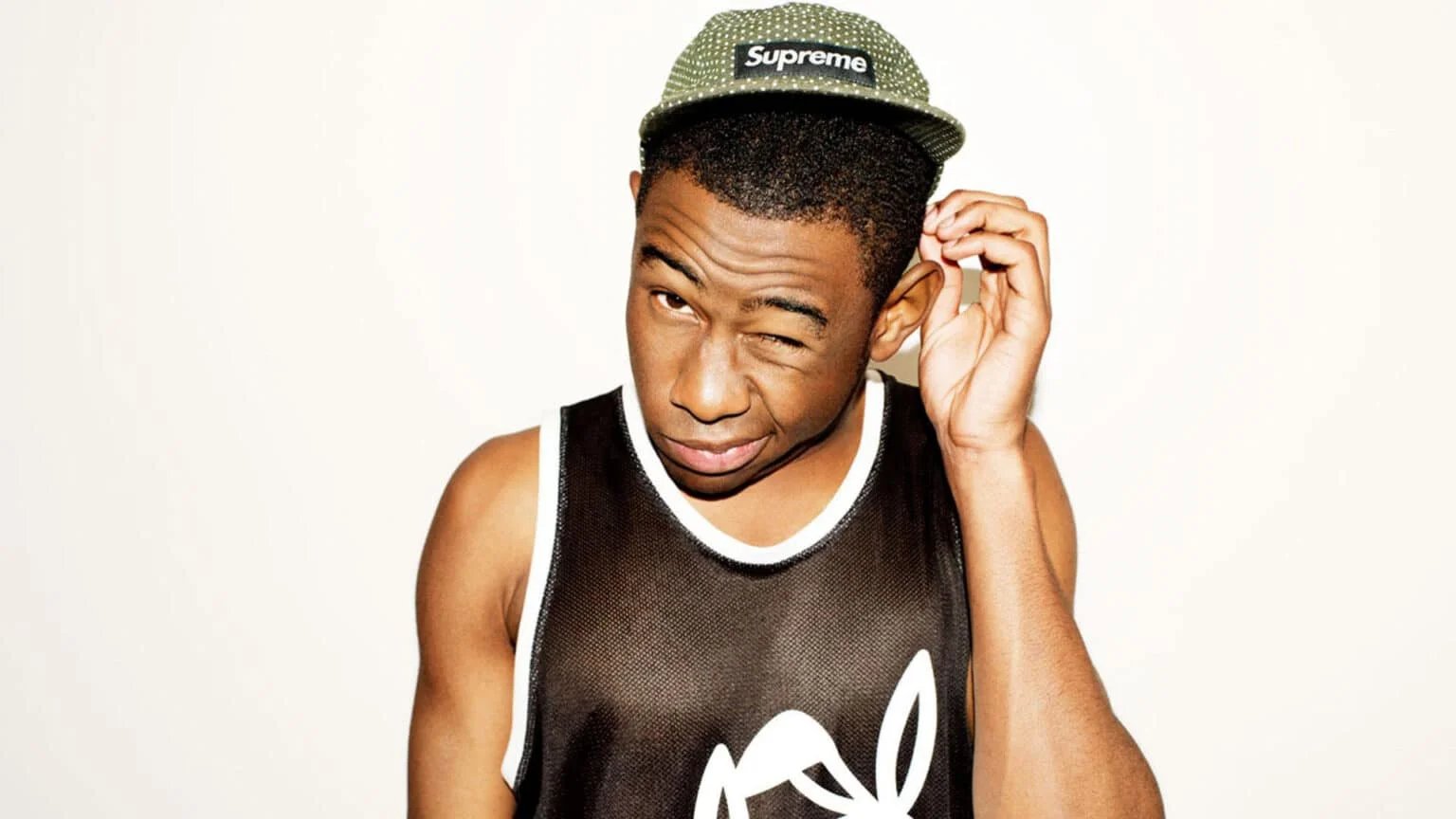 A person wearing a black tank top with a white outline and a graphic design is winking. They are also wearing a green cap with the word 'Supreme' on it, and are slightly scratching their head.