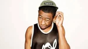 A person wearing a black tank top with a white outline and a graphic design is winking. They are also wearing a green cap with the word 'Supreme' on it, and are slightly scratching their head.
