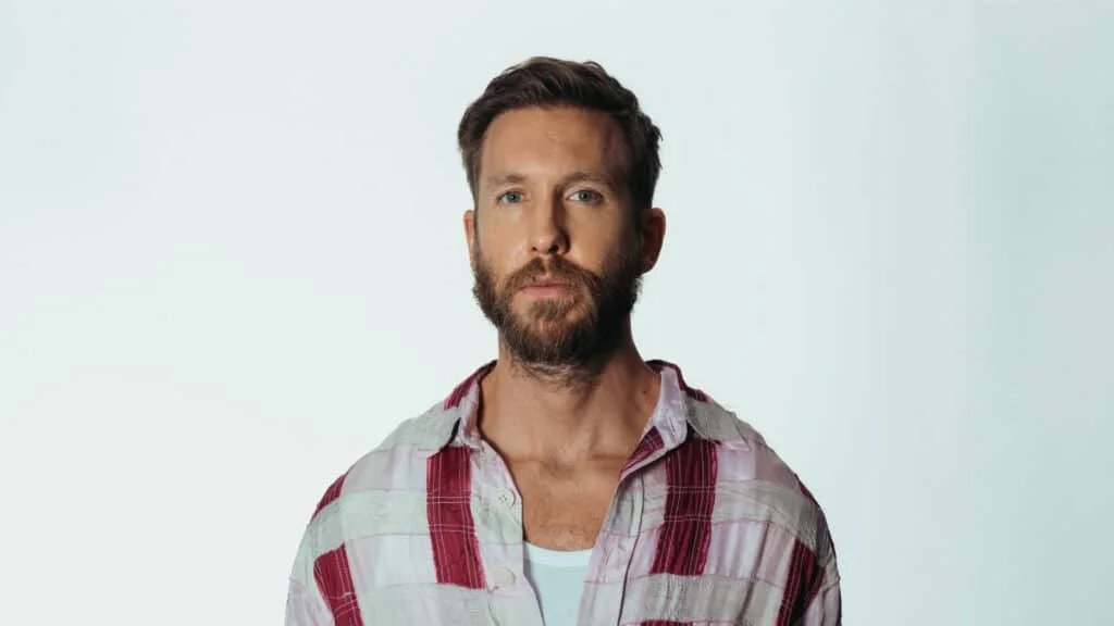 A man with a beard and short hair, wearing a plaid shirt and a white undershirt, standing against a plain white background.
