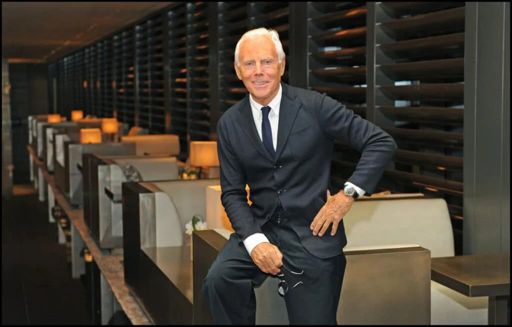 An older man with white hair wearing a dark suit and tie, leaning casually against a divider in a modern, dimly lit restaurant or lounge area, holding a pair of glasses in one hand.