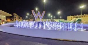 A night view of a fountain with illuminated water jets, featuring a large letter "W" in the background, surrounded by palm trees and bright streetlights.