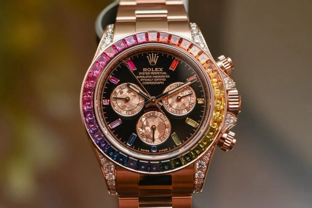 A luxurious wristwatch with a rose gold strap and a bezel adorned with multicolored gemstones. The black watch face features three sub-dials and diamond markers, with additional diamond embellishments on the case.