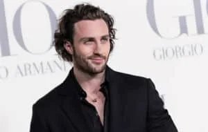 A man with wavy brown hair and a beard wearing a black suit, standing against a backdrop with Giorgio Armani branding.