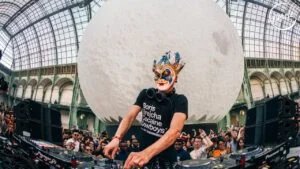 A DJ wearing a distinctive theatrical mask and a black T-shirt is performing in a large, glass-domed venue. A crowd of people is gathered around him, enjoying the music. The room is dominated by an enormous spherical object resembling a moon as a backdrop.
