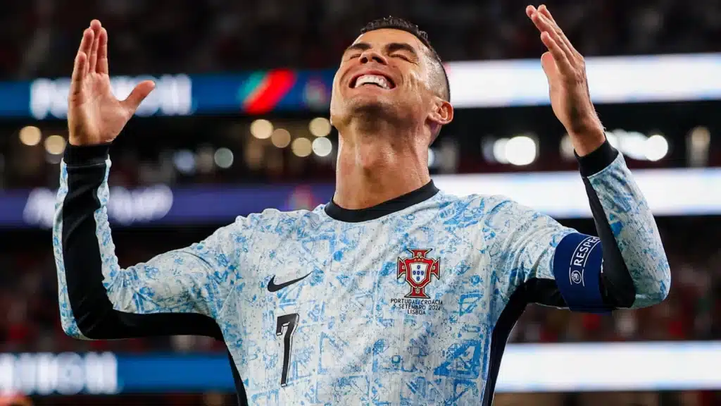 A soccer player wearing a light blue and white Portugal jersey, with the number 7 and a captain's armband, looks upwards with an expression of exasperation or frustration on his face. The background is blurred, suggesting a stadium setting.