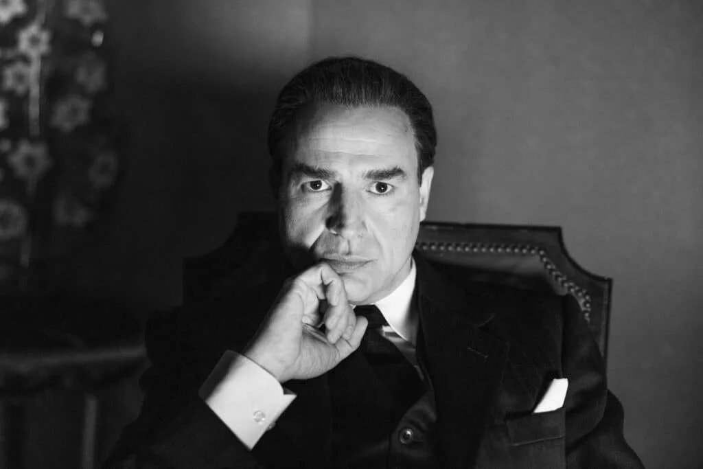 Black and white portrait of a man in a suit sitting in a chair, with his hand resting on his chin in a thoughtful pose.