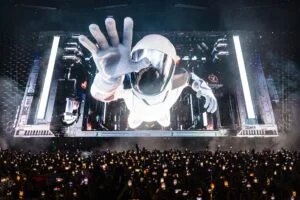 A large digital screen displays an astronaut reaching out with one hand, while a crowd of people holding up phones watches the scene below.