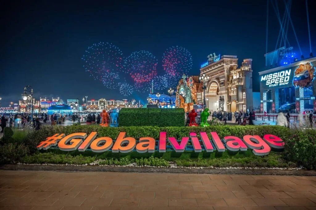 A vibrant nighttime scene at Global Village, featuring large illuminated letters spelling '#Globalvillage' in the foreground, surrounded by lush greenery. In the background, a bustling fairground with brightly lit buildings is visible, including a sign for 'Mission Speed Stunt Show.' Fireworks explode in the night sky, adding a festive ambiance.