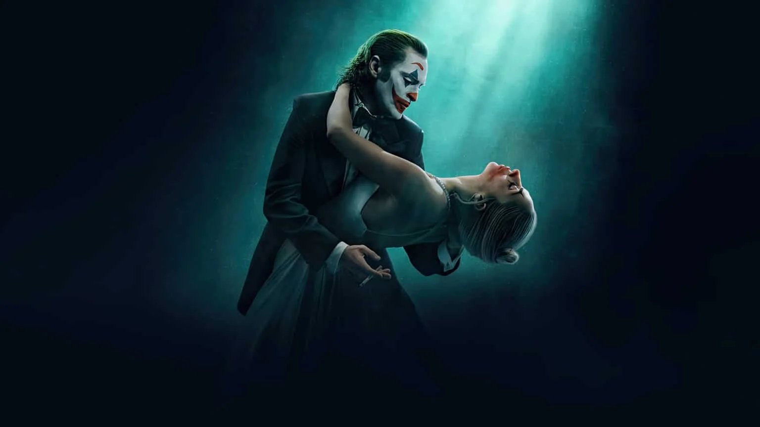 A person dressed in a dark suit with clown makeup holds another person wearing a light-colored dress and pearl necklace in a dance pose under a dramatic spotlight.