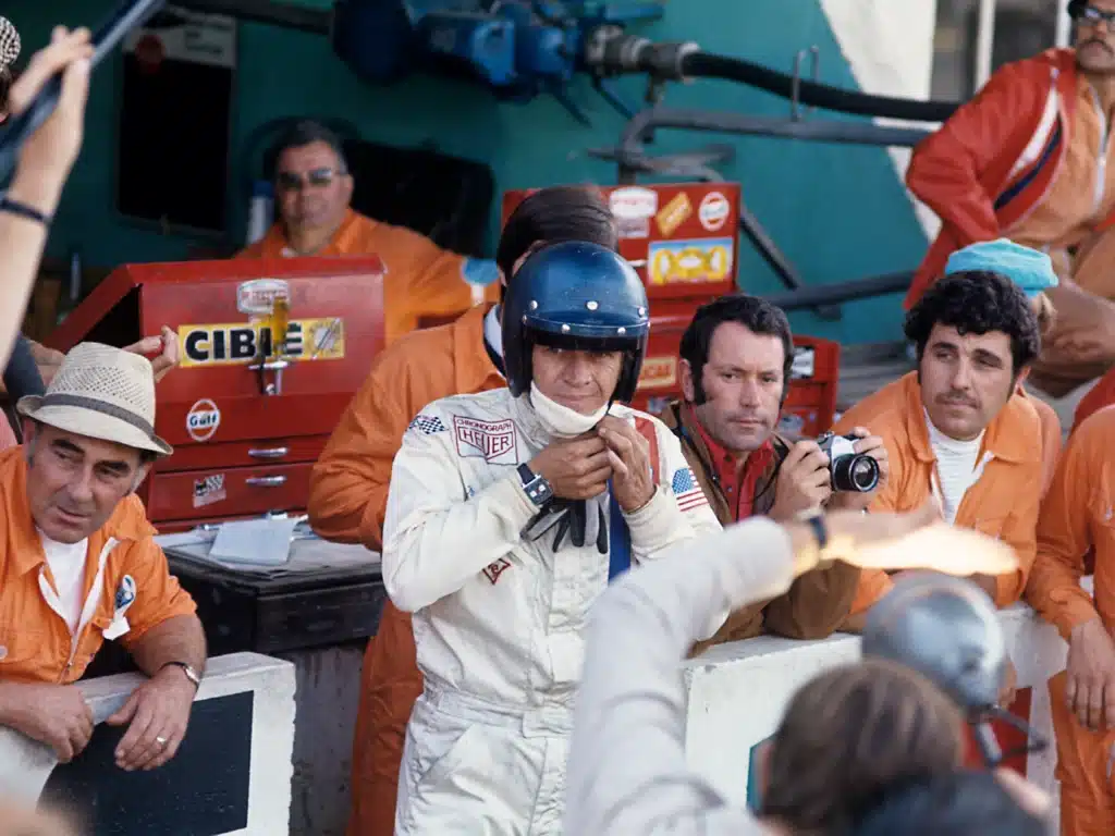 A race car driver in a white suit and blue helmet adjusts his chin strap while surrounded by a group of men, some in orange uniforms. One man holds a camera, capturing the moment in a pit stop environment with a backdrop of toolboxes and equipment.