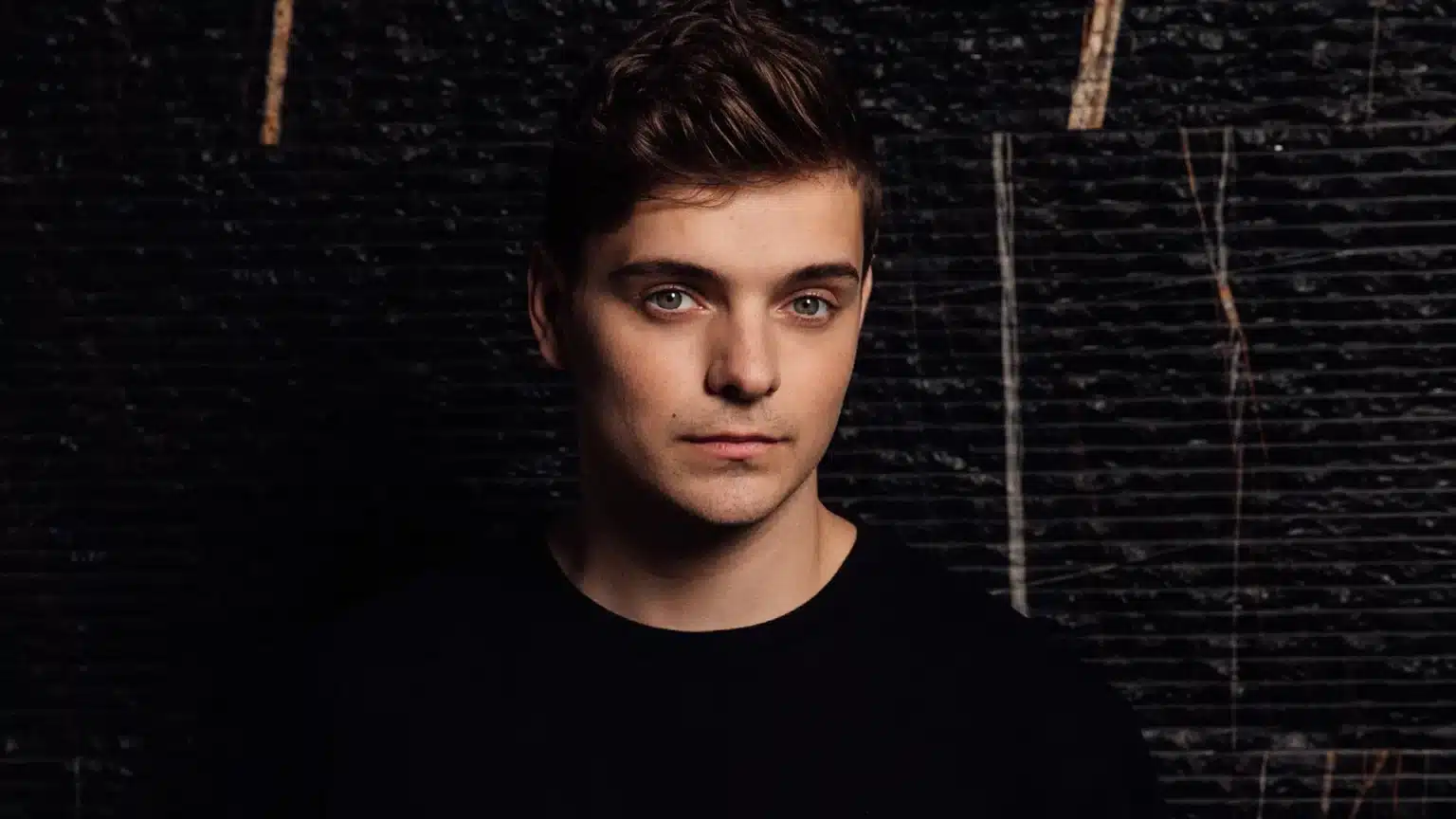 A young man with short brown hair and a serious expression, wearing a black shirt, stands against a dark, textured background.