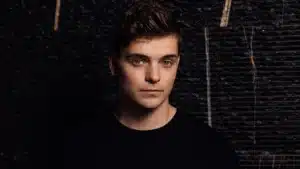 A young man with short brown hair and a serious expression, wearing a black shirt, stands against a dark, textured background.