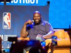 A smiling man with a beard holding a microphone, seated on a stage with a blue NBA-themed backdrop and an orange chair, surrounded by an audience taking photos.