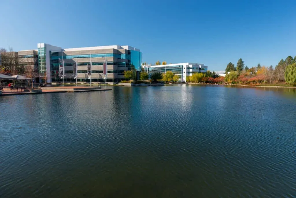 Modern office buildings with glass facades situated beside a calm body of water, surrounded by green and autumn-colored trees under a clear blue sky.