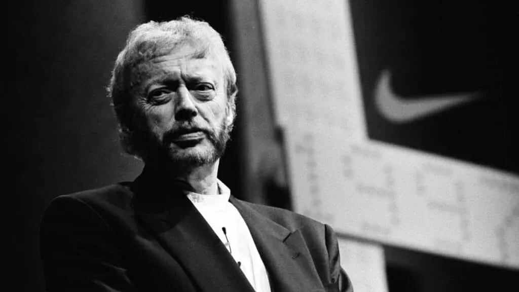 A black and white image of a man with a beard and suit standing in front of a background featuring the Nike logo.