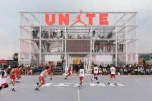 A basketball game in progress on a gray outdoor court with players in red and white jerseys, near a large structure with the word 'UNITE' and a logo in red at the top. Spectators are watching from the stands and the building's balconies.