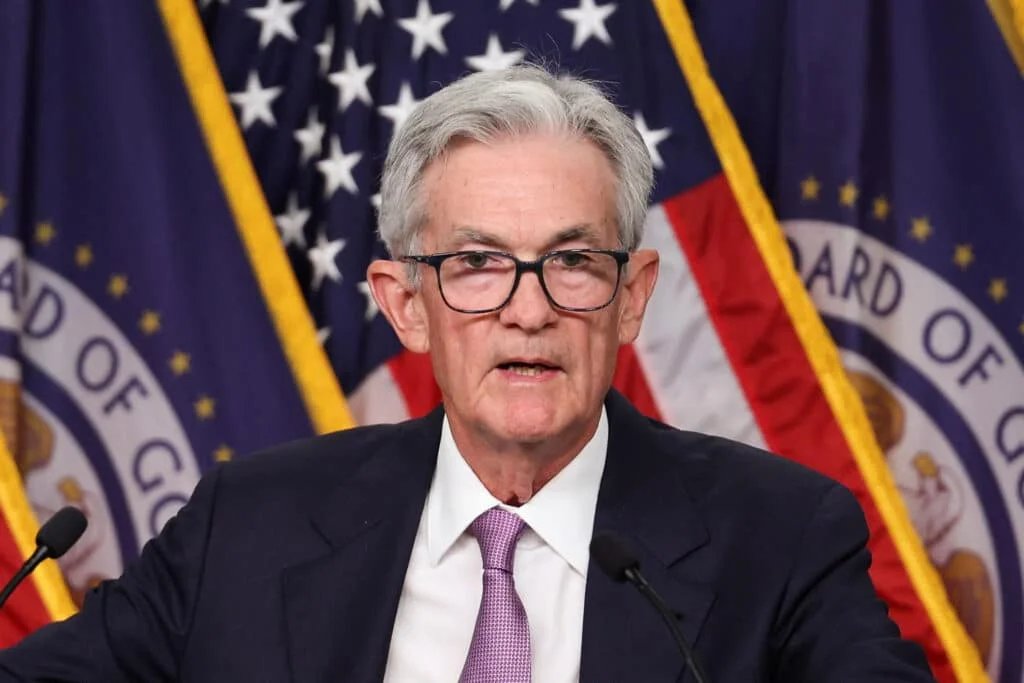 A man wearing glasses and a suit speaks at a podium with American flags and a board logo in the background.