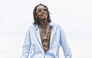 Man with dreadlocks wearing sunglasses, an open light blue shirt, and multiple necklaces, smiling and looking upward.