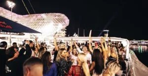 A lively group of people partying on a boat at night, with individuals raising their arms and holding drinks. The background features an illuminated, futuristic-looking structure with patterned lights, and reflections can be seen on the surrounding water.