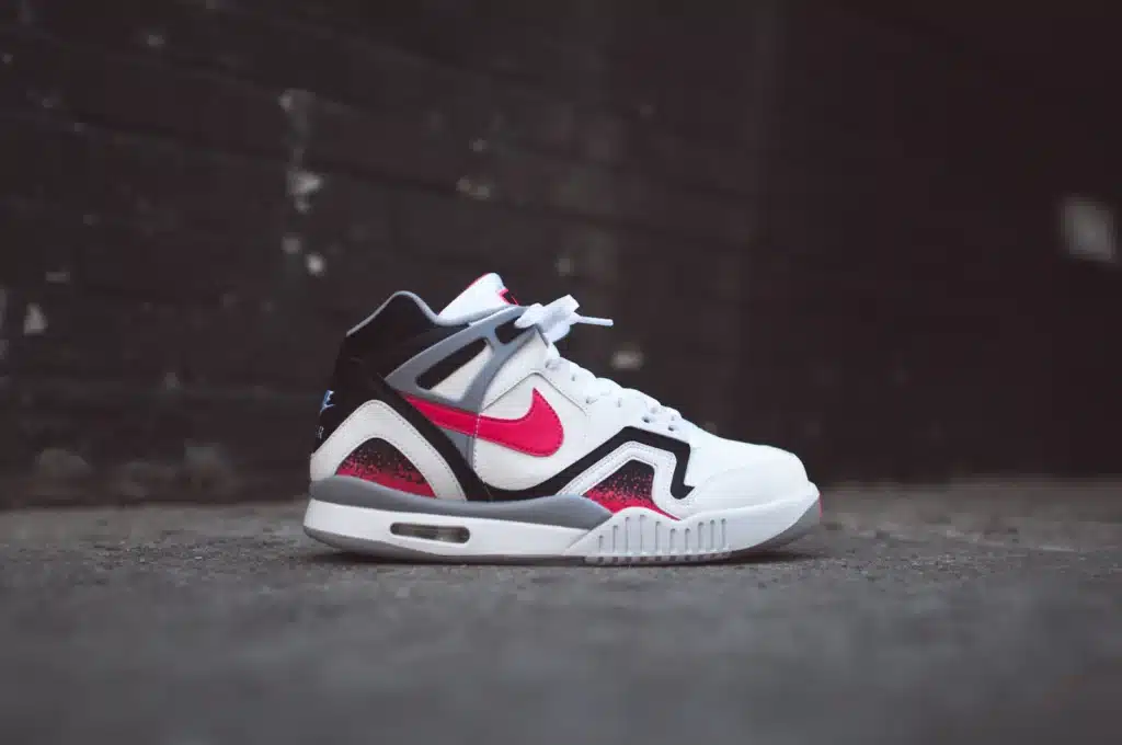 A side view of a white and black sneaker with red accents, featuring a prominent red swoosh logo, resting on a concrete surface against a dark, blurred background.