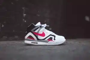 A side view of a white and black sneaker with red accents, featuring a prominent red swoosh logo, resting on a concrete surface against a dark, blurred background.