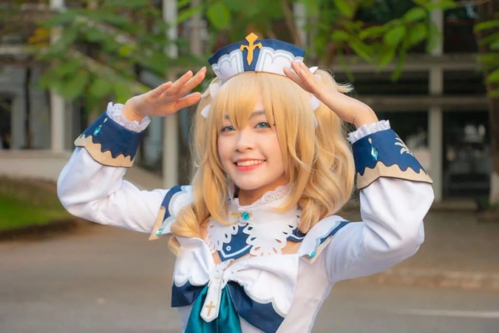 A person wearing a detailed blue and white costume with a matching headpiece, featuring blonde hair and a cheerful smile, posing outdoors with hands raised to their forehead.