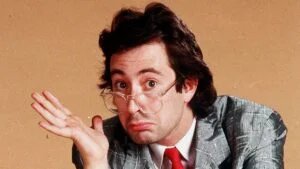 A person with thick-framed glasses and a humorous expression, shrugging while wearing a gray suit with a red tie against a neutral background.