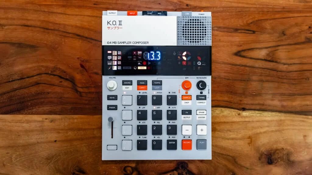 A K.O. II 64 MB sampler composer with a light gray body, black and orange buttons, and a digital display, placed on a wooden surface.
