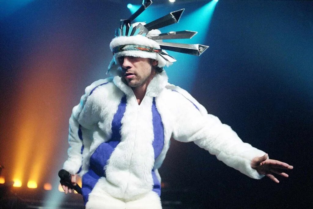 Person wearing a unique outfit with a white fur coat and a headpiece with metallic spikes, performing on stage under colorful lights.