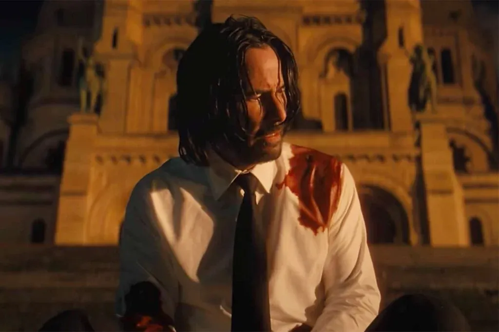 A man with shoulder-length dark hair, wearing a white shirt stained with blood on the shoulder and sleeve, sits in front of a lit-up, ornate building at night.