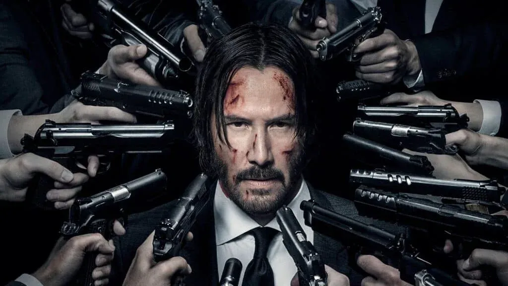 A man with a serious expression and scrapes on his face is surrounded by numerous hands pointing guns at him.