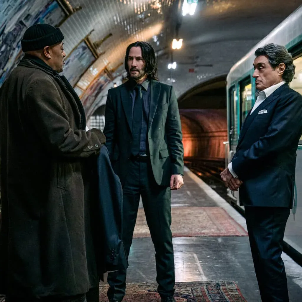 Three men stand in a dimly lit subway station; one in a dark coat and beanie, another with long hair in a dark suit, and the third in a suit with white shirt, each appearing engaged in conversation.