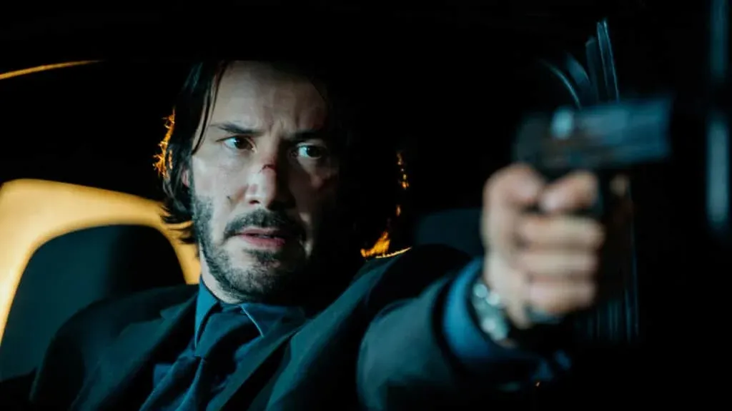 A man sitting in a dimly lit car interior, wearing a suit, holds a gun pointed forward.