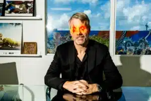 A man with glowing red and orange eyes sits at a glass table in an office setting, wearing a black suit. The background features colorful murals visible through the windows and several framed artworks on the wall.