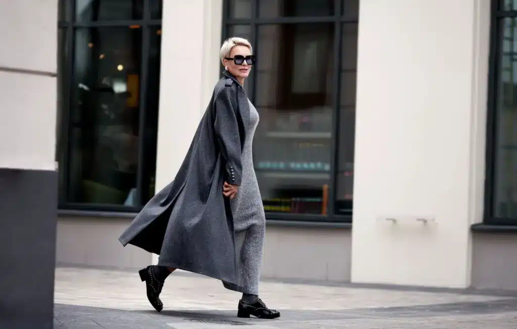 A person with short blonde hair, wearing a long gray coat and sunglasses, walks confidently on a city street, with large windows of a building in the background.