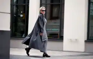 A person with short blonde hair, wearing a long gray coat and sunglasses, walks confidently on a city street, with large windows of a building in the background.