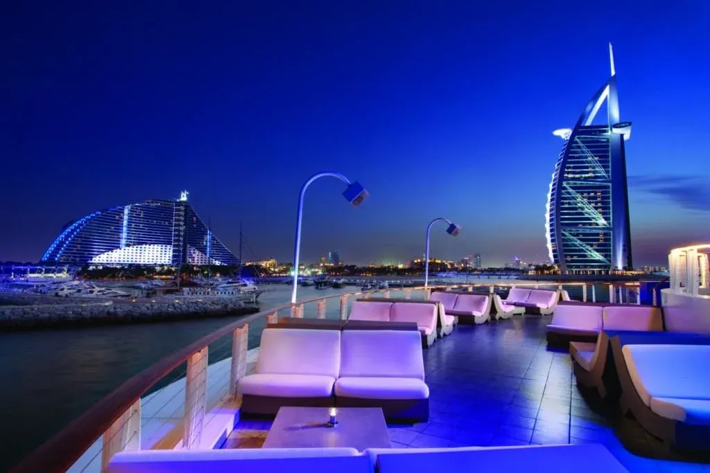 View of a waterfront location at night featuring modern seating and ambient lighting, with two iconic, illuminated buildings in the background.