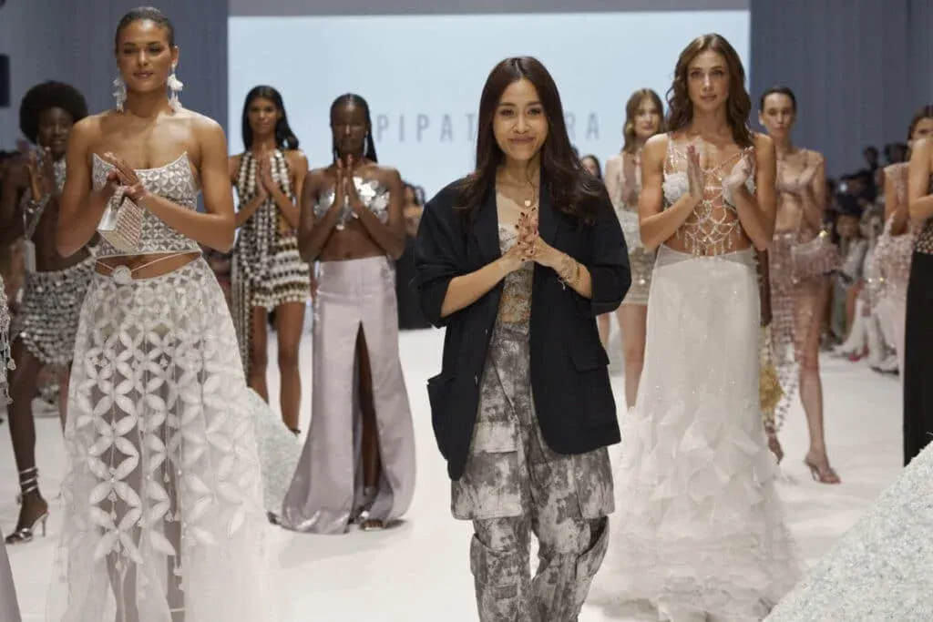 A fashion designer walks on the runway surrounded by models wearing intricate, avant-garde outfits, following a fashion show presentation.