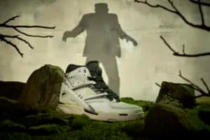 A sneaker designed with a Frankenstein theme is placed on a mossy forest floor, with a silhouette of a Frankenstein-like figure on the wall in the background.