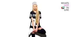 A person with long blonde hair wearing a gray beanie, a black oversized T-shirt with white stripes and text, knee-high black socks, and white sneakers, standing on one leg while grimacing and winking. "Whats Hot in UAE" text with an abstract flag design is on the right side of the image.