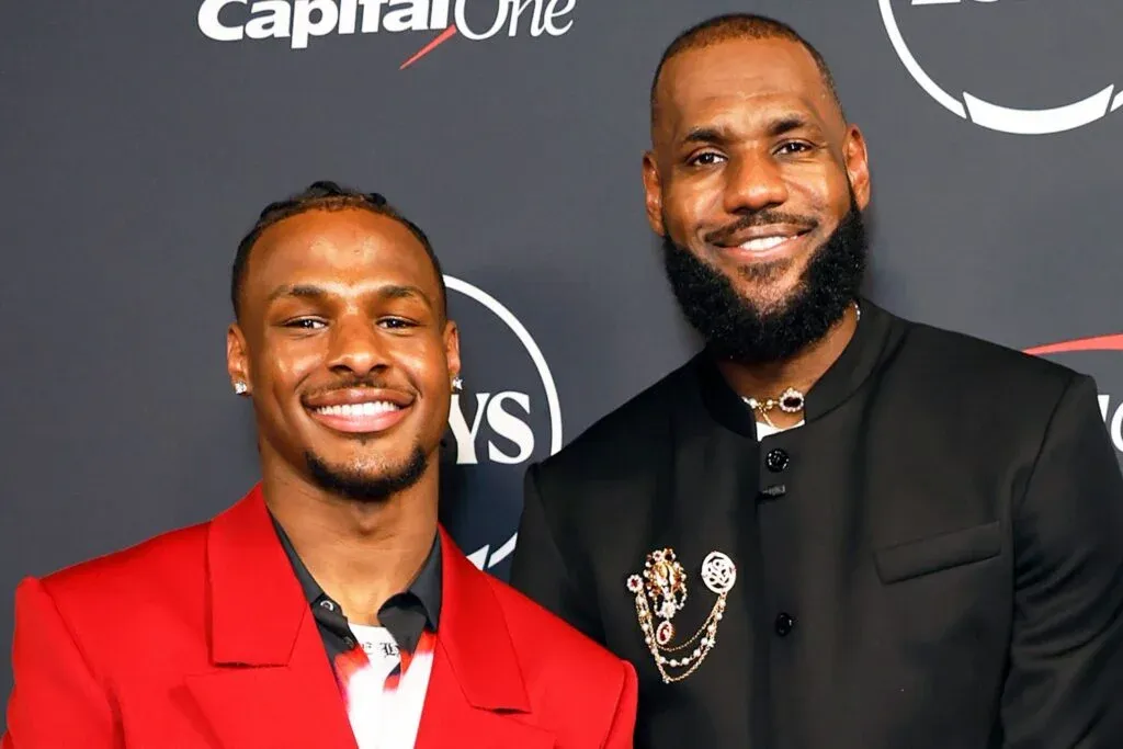 Two men smiling, one wearing a red suit jacket and the other in a black jacket with decorative pins, stand together against a backdrop with logos including 'Capital One' and 'ESPYS.'
