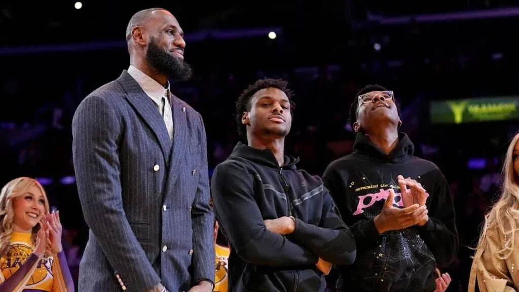 Three men standing together, with the one on the left wearing a suit, the middle one wearing a black jacket, and the one on the right wearing a black hoodie with graphics, all against a background of a basketball court.