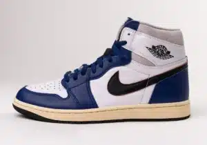 A high-top sneaker featuring a white and blue leather upper, black Nike swoosh, blue laces, and beige midsole, with a logo on the ankle area.