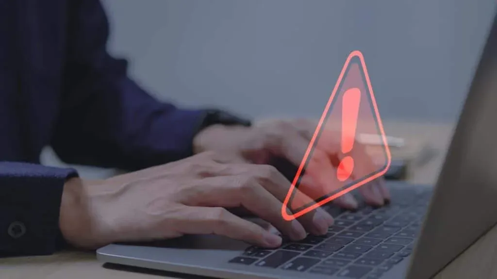 A person typing on a laptop keyboard with a red warning triangle and exclamation mark symbol appearing in the foreground, indicating a potential alert or error.