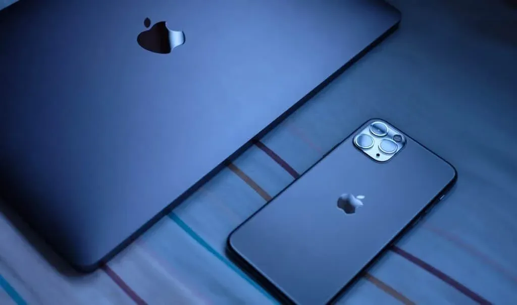 A MacBook and an iPhone with a triple-camera setup, both featuring the Apple logo, placed next to each other on a bed with a striped bedsheet under soft lighting.