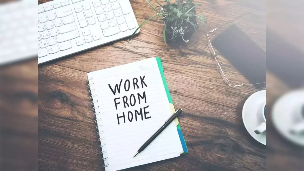 A desk setup with a notebook displaying the words 'WORK FROM HOME' next to a pen, a keyboard, a small plant, a pair of glasses, a smartphone, and a cup of coffee.